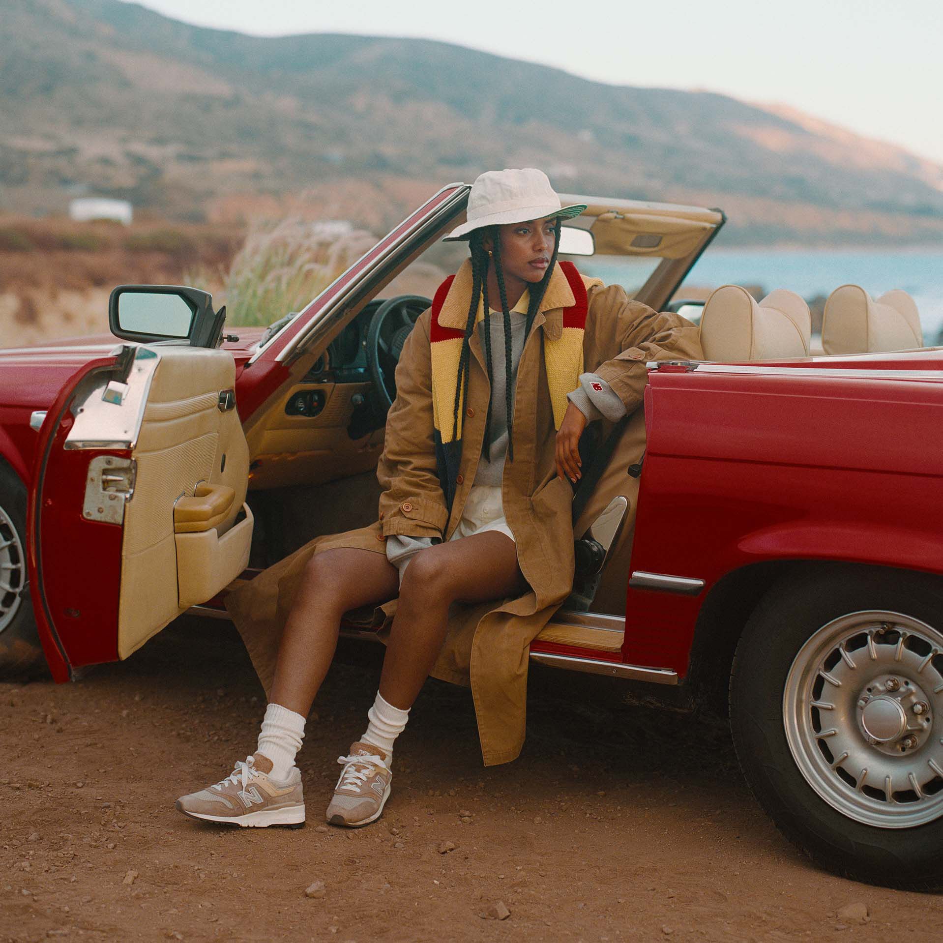 A woman sitting in a red car with the door open with a beach landscape behind her. 