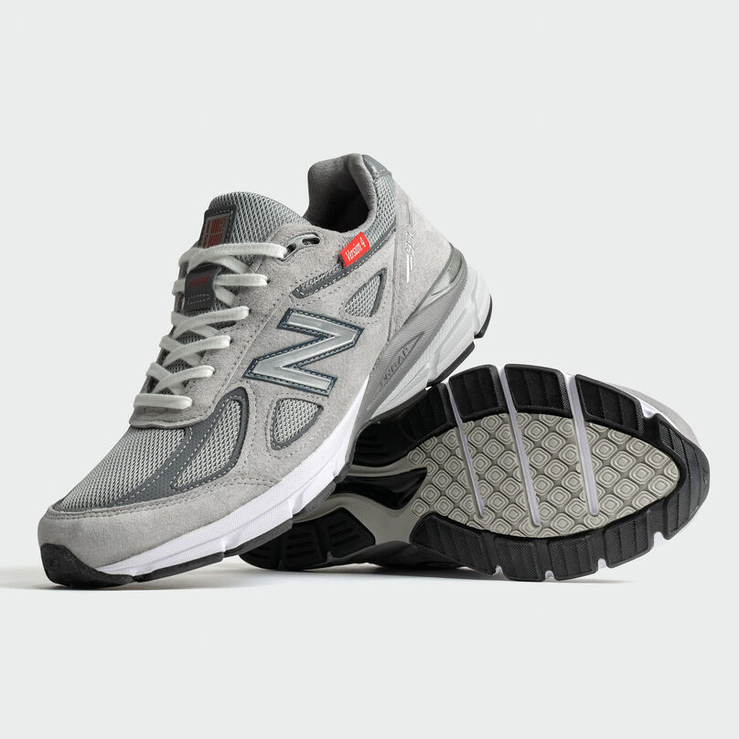 Pair of grey 990v4 sneakers on a grey studio background with one shoe propped on the other.