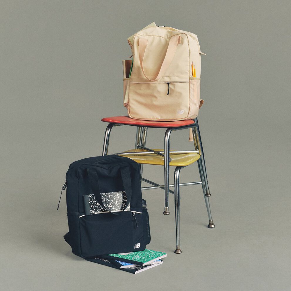 Studio image of a pair of cream-colored New Balance sneakers placed on a dark wooden folding chair, with a second matching chair folded and leaned behind it.