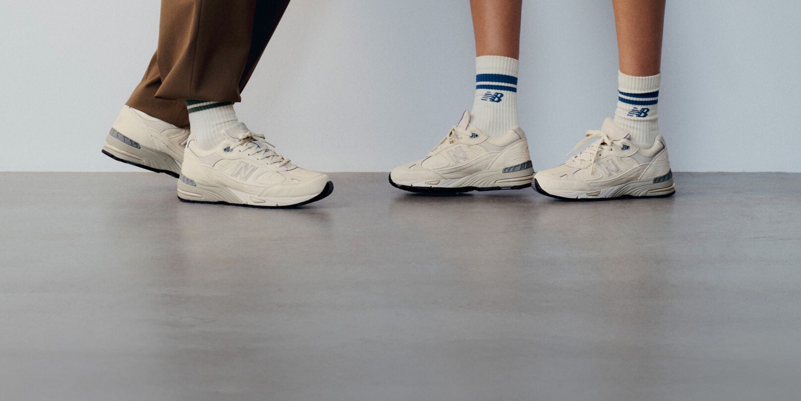 Knee down view of a man and woman wearing New Balance Made in UK sneakers standing on a concrete floor against a grey wall. 