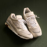 Knee down view of a man wearing a pair of New Balance Made in UK sneakers standing on a rustic wooden floor with smashed blue and white ceramic crockery all over the ground and white cabinets in the background. 