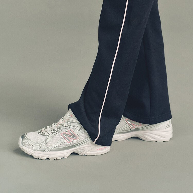 Close-up studio image of the lower legs of someone wearing New Balance sneakers against a plain light gray background.
