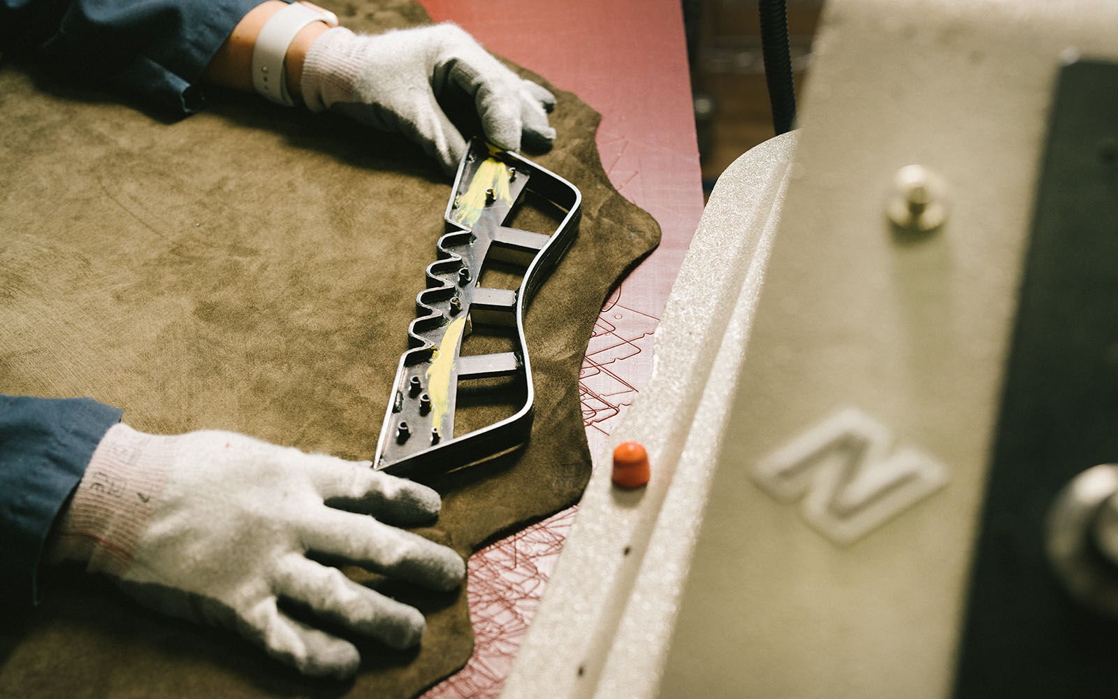 A pair of hands laying a tool over a piece of suede in a factory setting. 