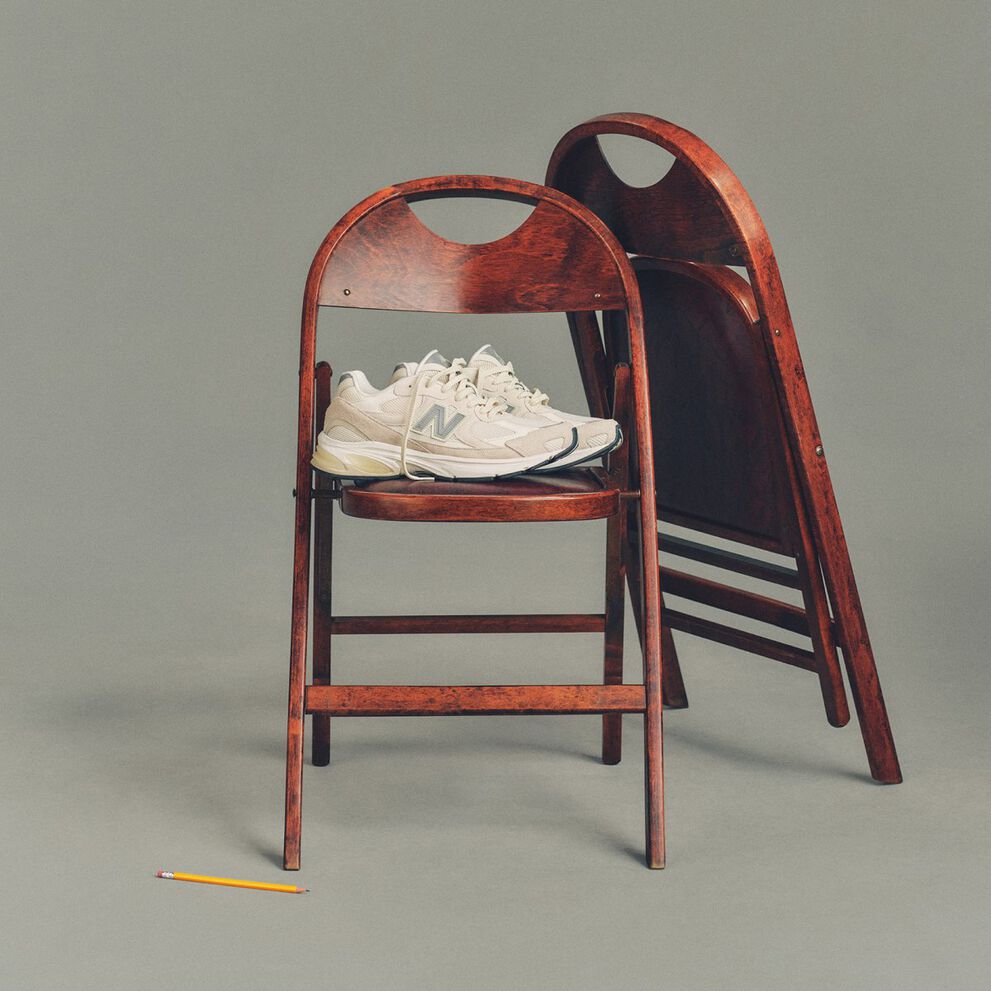 Studio image of a pair of cream-colored New Balance sneakers placed on a dark wooden folding chair, with a second matching chair folded and leaned behind it.