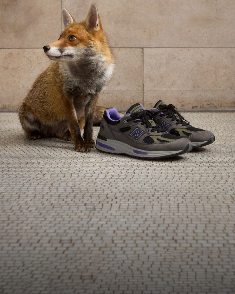 A pair of New Balance Made in UK sneakers on a white tiled floor with stone walls in the background next to a sitting fox. 