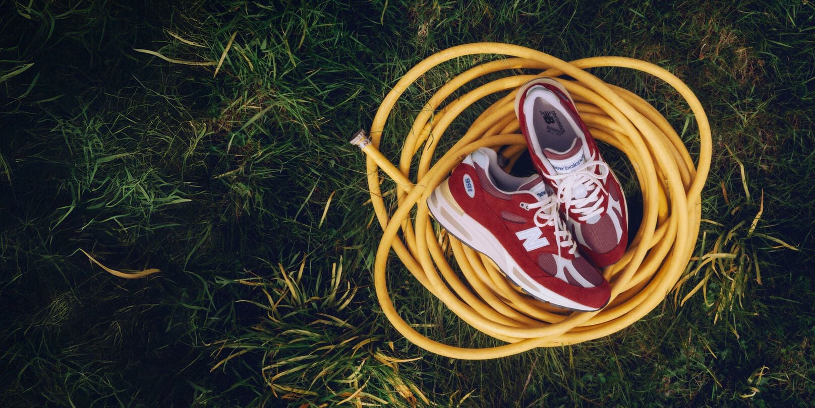 A pair of New Balance Made in UK sneakers sitting on a yellow coiled hose on top of a lush grass lawn. 