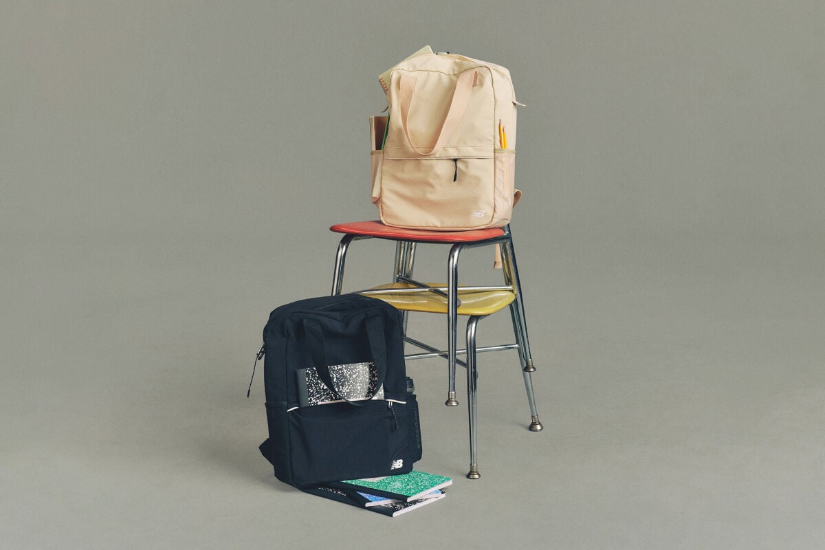 Studio image of a pair of cream-colored New Balance sneakers placed on a dark wooden folding chair, with a second matching chair folded and leaned behind it.